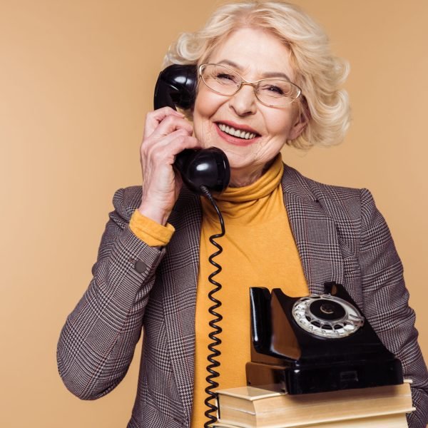 happy fashionable senior woman in eyeglasses talking on rotary phone and holding stack of books