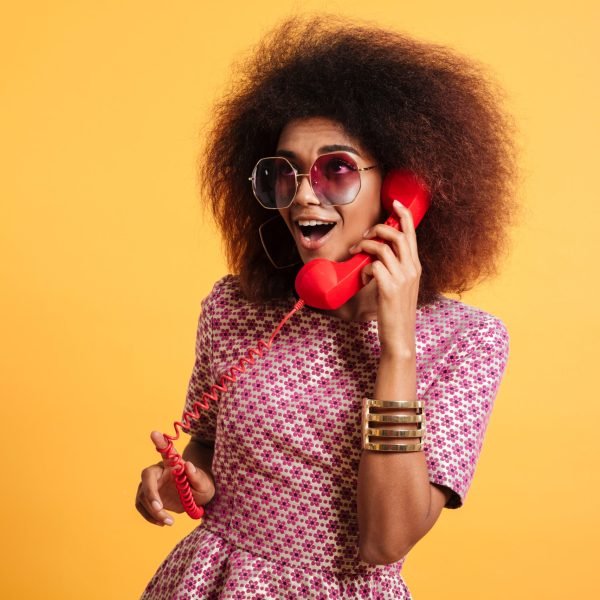 Portrait of an excited pretty afro american woman in retro style clothes talking on telephone isolated over yellow background